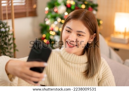 Young asian woman wearing sweater and smiling while sitting to relaxation and using smartphone for taking a photo in holiday on sofa in cozy living room of christmas decoration at home.