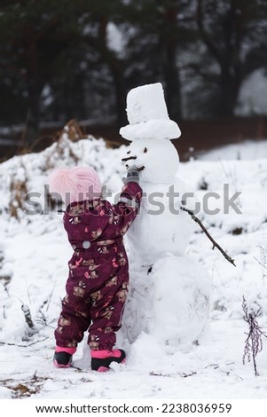 A four-year-old girl builds a snowman in the winter in the forest, around a snowdrift. Vertical orientation.