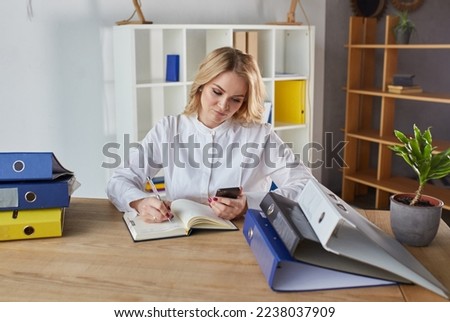 The girl at the work table with a notebook and a smartphone. A young woman with short blonde hair in an office white shirt surfing the Internet from the phone. Close up.
