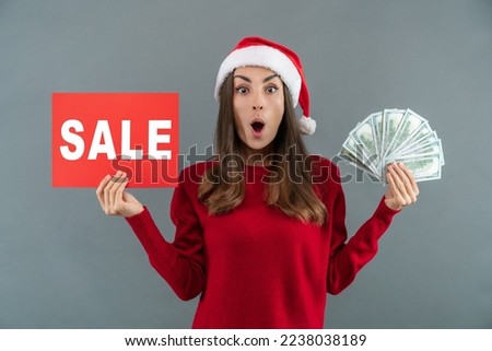 Studio shot of excited young woman in Santa's hat posing over grey background with Sale banner and dollar banknotes fan in hands. Holiday shopping concept.