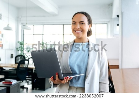 Photo of beautiful successful brazilian or latino business woman, in elegant clothes, seo, top manager, programmer, stands in a modern office, holding an open laptop in hands, looks at camera, smiling