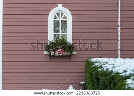A red exterior house wall of clapboard.  A large window with a curved or arched top. There's a large flower box filled with greenery and red Christmas bows on the wall. A green hedge has snow below.