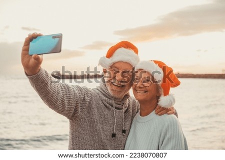 Two old happy seniors wearing christmas hats at the beach taking a selfie of them smiling and having fun with the sunset at the background at evening. Cute couple of old persons looking at the camera
