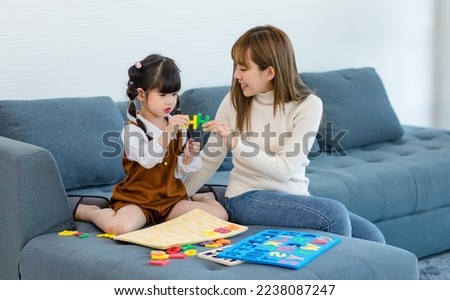 Millennial Asian happy cheerful little cute preschooler daughter girl playing learning alphabets letters jigsaw toy on sofa in living room while young teenager mother nanny helping teaching supporting