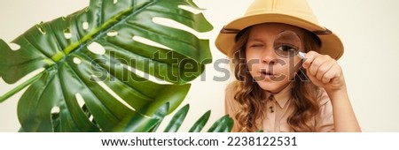 Child discoverer. Traveler child. Young explorer. A little girl in a safari hat with a magnifying glass looks carefully through the green palm leaves. Concept: search for adventure and treasure. Royalty-Free Stock Photo #2238122531
