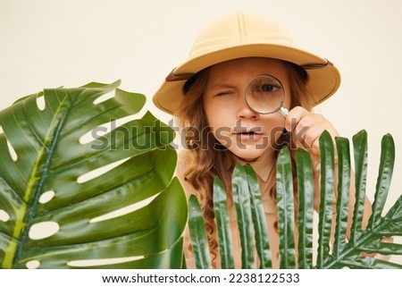 Child discoverer. Traveler child. Young explorer. A little girl in a safari hat with a magnifying glass looks carefully through the green palm leaves. Concept: search for adventure and treasure. Royalty-Free Stock Photo #2238122533
