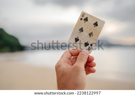 Old playing card six of spades in a man's hand against the backdrop of a tropical landscape.