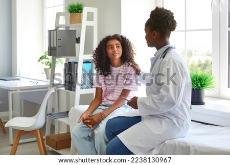Friendly female pediatrician touches shoulder of teenage girl and informs her that there is no threat to her health. African American doctor and her preteen patient are talking in hospital office. Royalty-Free Stock Photo #2238130967