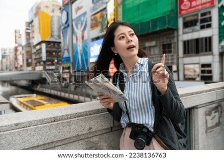 Asian female tourist finger pointing at distance while consulting a map on bridge over doutonbori river to find where she is in shinsaibashi suji Osaka japan