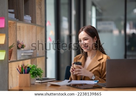 Beautiful woman holding a coffee cup and using a laptop while sitting at her working place in an office.