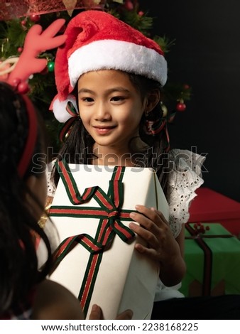 A pretty little Indonesian Chinese girl with Santa hat and white dress looks happy as she is receiving a Christmas gift from a friend. Isolated in Black Background.