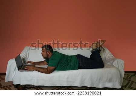 serious busy beard man lying on white sofa and working with mobile cell phone and laptop. pink wall background. picture frame. white mock up. 