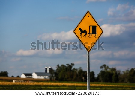 Yellow road sign for Amish buggy crossing and traffic with farm out of focus in background.
