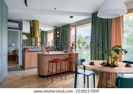Interior design of harmonized dining room with kitchen, round table, green chairs, wooden kitchen island, gold cooker hood, brastools, vase with leaves and personal accessories. Home decor. Template. Royalty-Free Stock Photo #2238217009