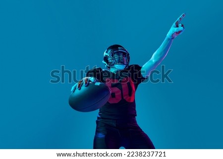 Sportive, strong man, american football player in sports team uniform and protective helmet isolated over blue background in neon light. Power, energy, achievements, skills and ad