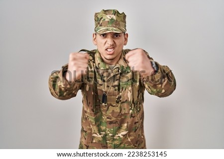 Young arab man wearing camouflage army uniform angry and mad raising fists frustrated and furious while shouting with anger. rage and aggressive concept. 
