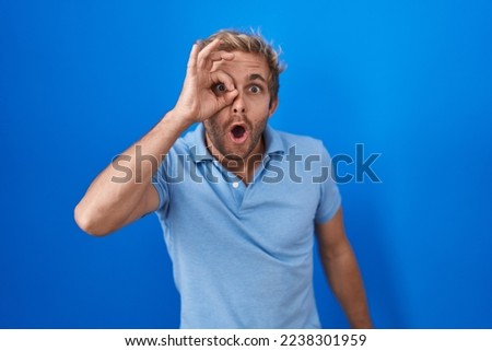 Caucasian man standing over blue background doing ok gesture shocked with surprised face, eye looking through fingers. unbelieving expression. 