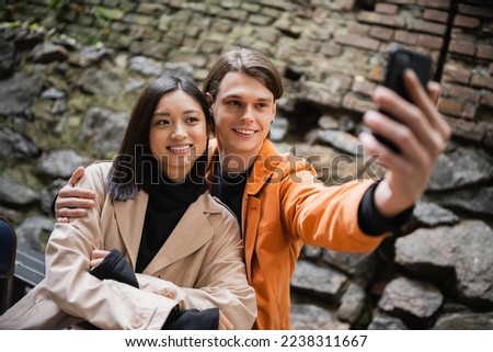 Smiling man hugging asian girlfriend while taking selfie on smartphone on terrace of cafe
