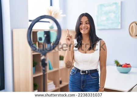 Brunette woman recording vlog tutorial with smartphone at home smiling with an idea or question pointing finger up with happy face, number one 