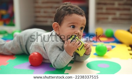 Adorable hispanic baby sucking car toy lying on floor at kindergarten Royalty-Free Stock Photo #2238337515