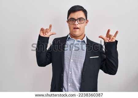 Young hispanic man with down syndrome wearing business style shouting with expression doing rock symbol with hands up. music star. heavy concept. 
