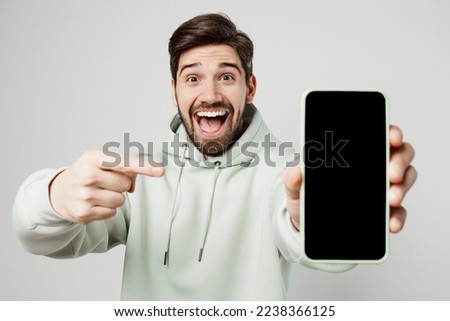 Young amazed fun impressed happy man wear mint hoody hold in hand use point index finger on mobile cell phone with blank screen workspace area isolated on plain solid white background studio portrait