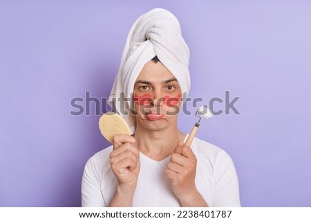 Portrait of sad man with patches under eyes wearing white T-shirt and being wrapped in towel standing isolated over purple background, holding sponge and massage roller.
