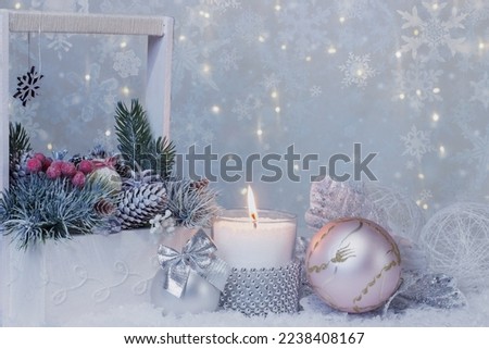 A white wooden basket with vintage lace and a bouquet of fir branches and cones, a burning candle in a decorative candlestick and two Christmas balls on a light blurred background of snowflakes 