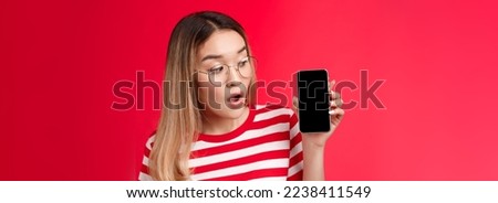 Close-up wondered surprised attractive asian blond girl wear glasses hold smartphone looking impressed amused phone screen, open mouth interesting app telephone display, stand red background.