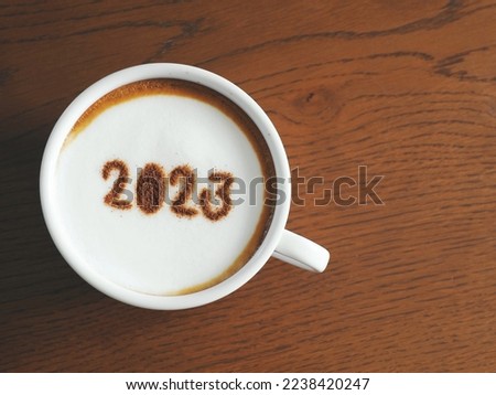 Welcome 2023 theme coffee cup with number 2023 on frothy surface flat lay on wooden table background. Happy new year 2023, holidays food art concept, food and drink trends. (close up, top view)