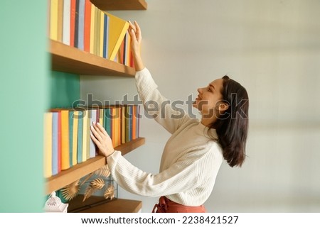 Beautiful young woman taking book from shelf in home library