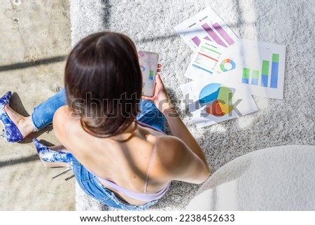 Overhead view of girl studying image of business charts on phone with charts on floor