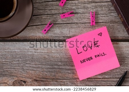 Love never fails, handwritten biblical verse on pink note with cup of coffee and holy bible book on wooden background. Top table view.