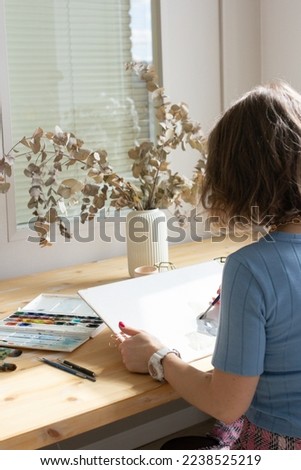 Young woman painting a picture with watercolor at wooden table, aquarelle process, girl sitting at the table and create picture