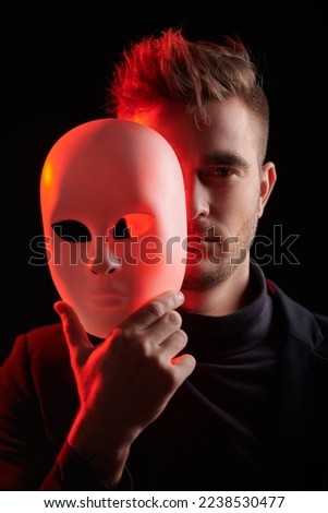 People and life roles. Portrait of a man in black suit who hides his face behind a mask. Black studio background with red light. Psychological art portrait. Royalty-Free Stock Photo #2238530477