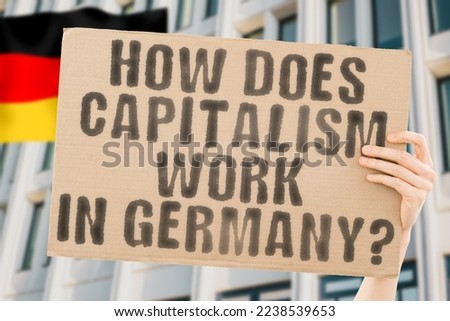 The question " How does capitalism work in Germany? " is on a banner in men's hands with blurred background. Person. Banking. Dollar. Market. Cash. Coin. Crisis. Exchange. Profit. Rich. Savings. Euro