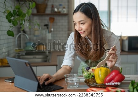 Asian housewife cutting fresh vegetables mixing salad ingredients cooking tasty vegan meal look search online blogger recipe notebook stand kitchen indoors