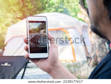 Handsome man taking photo of a camping site in a sunny summer forest with his mobile phone. Travel and camping concept.