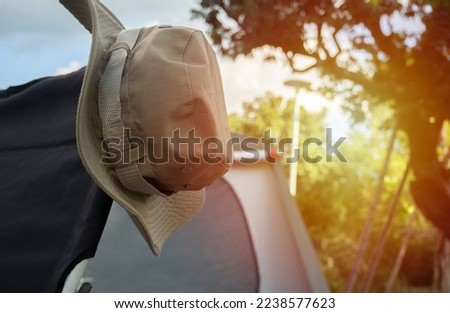 Clopped picture of hiking hat hanging on a camping chair with the background is a blurred image of the camping site. Camping adventure concept