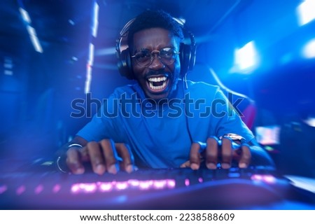 Happy American African Professional gamer, winner rejoices in victory of online games tournaments pc computer with headphones, Blurred red and blue background.