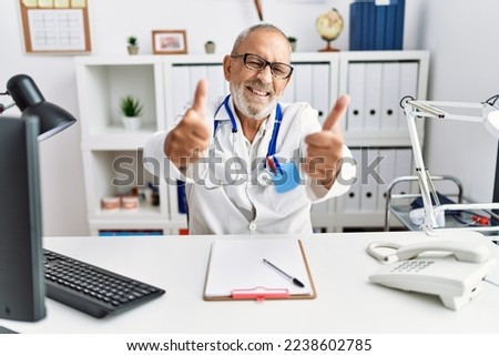 Mature doctor man at the clinic approving doing positive gesture with hand, thumbs up smiling and happy for success. winner gesture. 