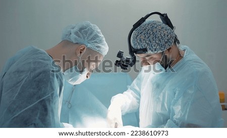 Two surgeons busy at work . Action . Two men in blue lab coats are painstakingly working on a complex operation , using various medical accessories like microscopic glasses