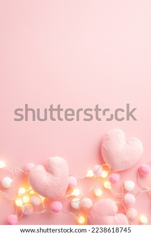 Valentine's Day concept. Top view vertical photo of fluffy heart shaped pillows light bulb garland and soft pompons on isolated pastel pink background with empty space