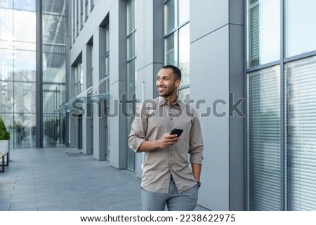 Smiling hispanic man walking down street near modern office building, freelancer businessman looking away holding mobile phone. Royalty-Free Stock Photo #2238622975