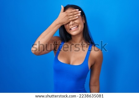 Hispanic woman standing over blue background smiling and laughing with hand on face covering eyes for surprise. blind concept. 