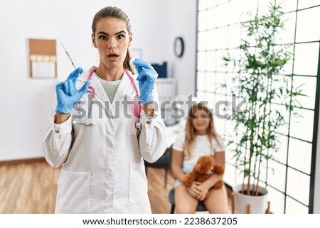 Young doctor woman putting vaccine to little girl in shock face, looking skeptical and sarcastic, surprised with open mouth 