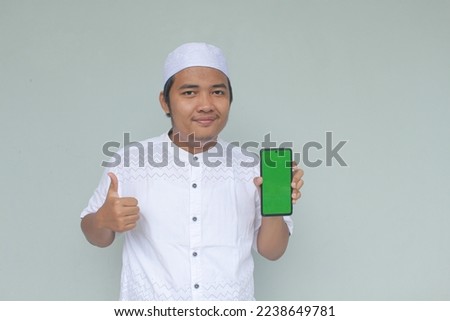 Smiling young Asian Muslim man give ok hand sign with confident gestures, while showing copy space on his phone, isolated by gray background.