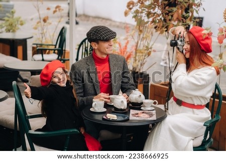 A stylish family of three is sitting at a table outside in a cafe and drinking coffee. Dad, mom and daughter in the autumn city