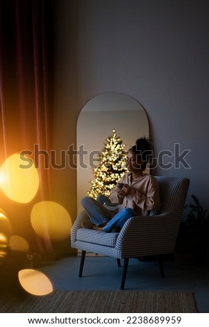 Merry Christmas and Happy Holidays! Young african american woman sitting in an armchair and drinking coffee by the Christmas tree in a cozy living room at home                              