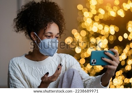 Merry Christmas and Happy Holidays! Young african american woman in wearing face mask video chatting on a smartphone on by the Christmas tree in the cozy living room of the house                      
